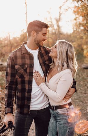 Engaged couple smiling at each other in autumn forest, woman showing engagement ring in warm sunlight.