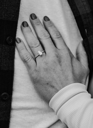 Black and white close-up of woman's hand with oval halo engagement ring and dark painted nails.