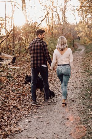 Couple walking hand in hand with black dog on a leaf-covered autumn forest trail at golden hour.