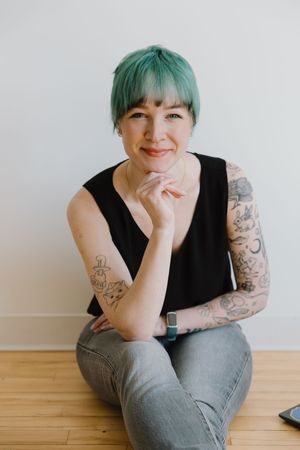 Tattooed woman with teal hair sitting on floor in black sleeveless top and jeans, smiling warmly.