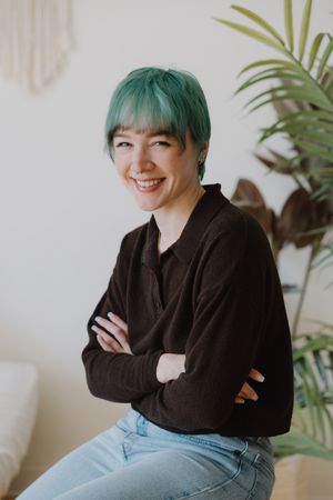 Smiling woman with short teal hair and crossed arms wearing a dark shirt and jeans indoors.