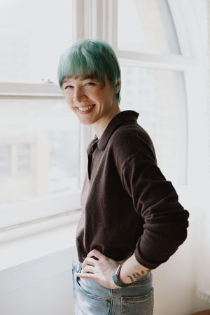 Cheerful woman with green hair in a dark sweater and jeans posing by a bright window.