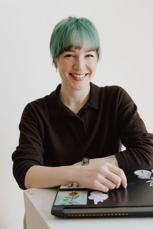 Smiling woman with short green hair leaning on a laptop at a white table indoors.