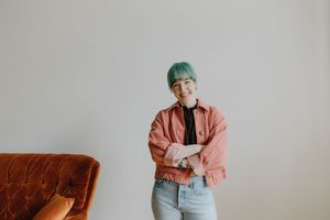 Happy woman with teal hair wearing a pink jacket and jeans standing near an orange chair.