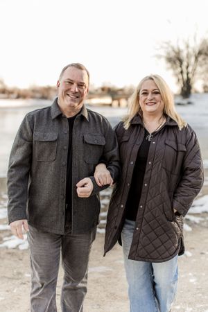 Smiling middle-aged couple in winter jackets pose together outdoors near a frozen lake at golden hour.
