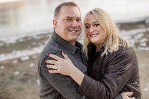 Happy couple hugging closely outdoors in winter, blonde woman wearing engagement ring smiling at camera.
