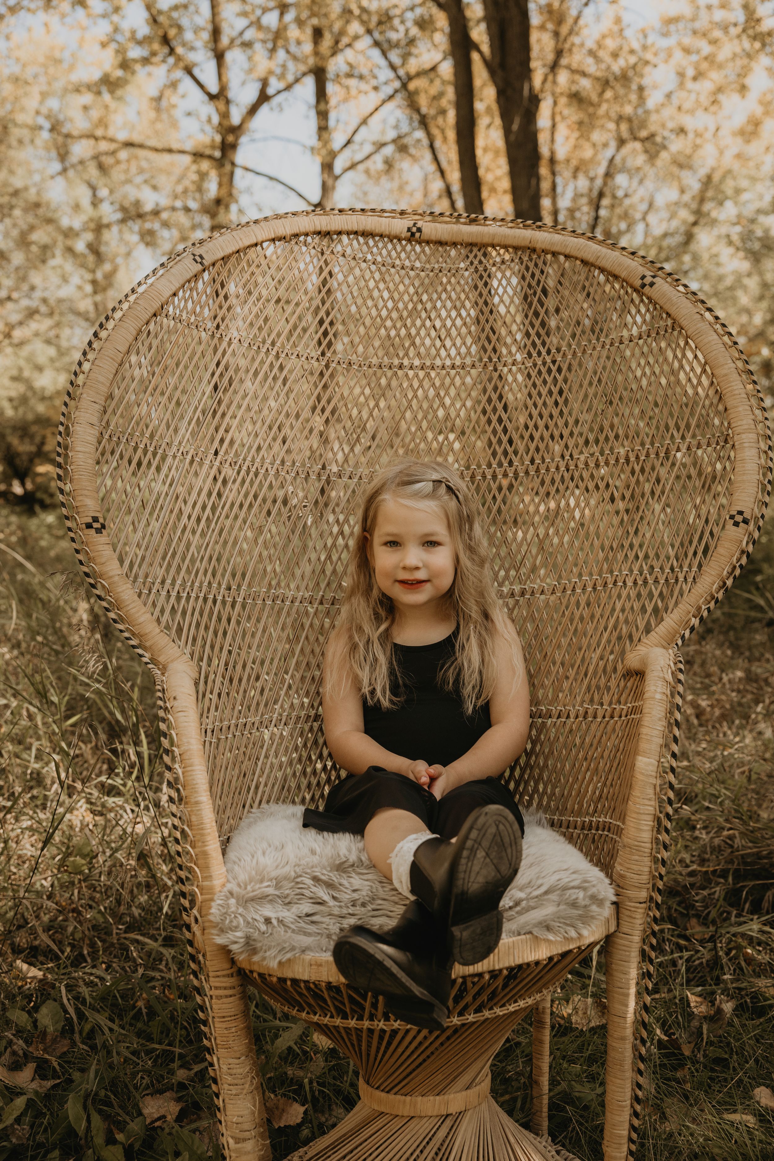 Cute blonde little girl sitting in large wicker peacock chair outdoors in autumn setting.