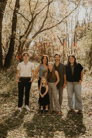 Family of six smiling together in a sunlit autumn forest with colorful fall foliage background.