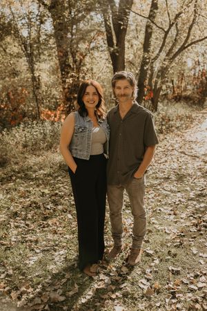 Smiling couple standing together in autumn forest with colorful fall leaves on the ground.