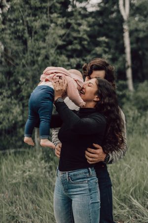 Happy mother lifting and kissing her baby girl in a lush green outdoor setting.