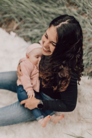 Smiling mother sitting on sandy ground holding her baby girl in a pink outfit.