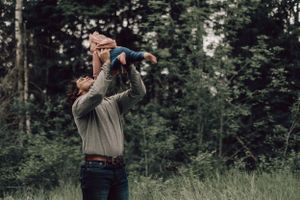 Father lifting baby in the air outdoors in a lush green forest setting.