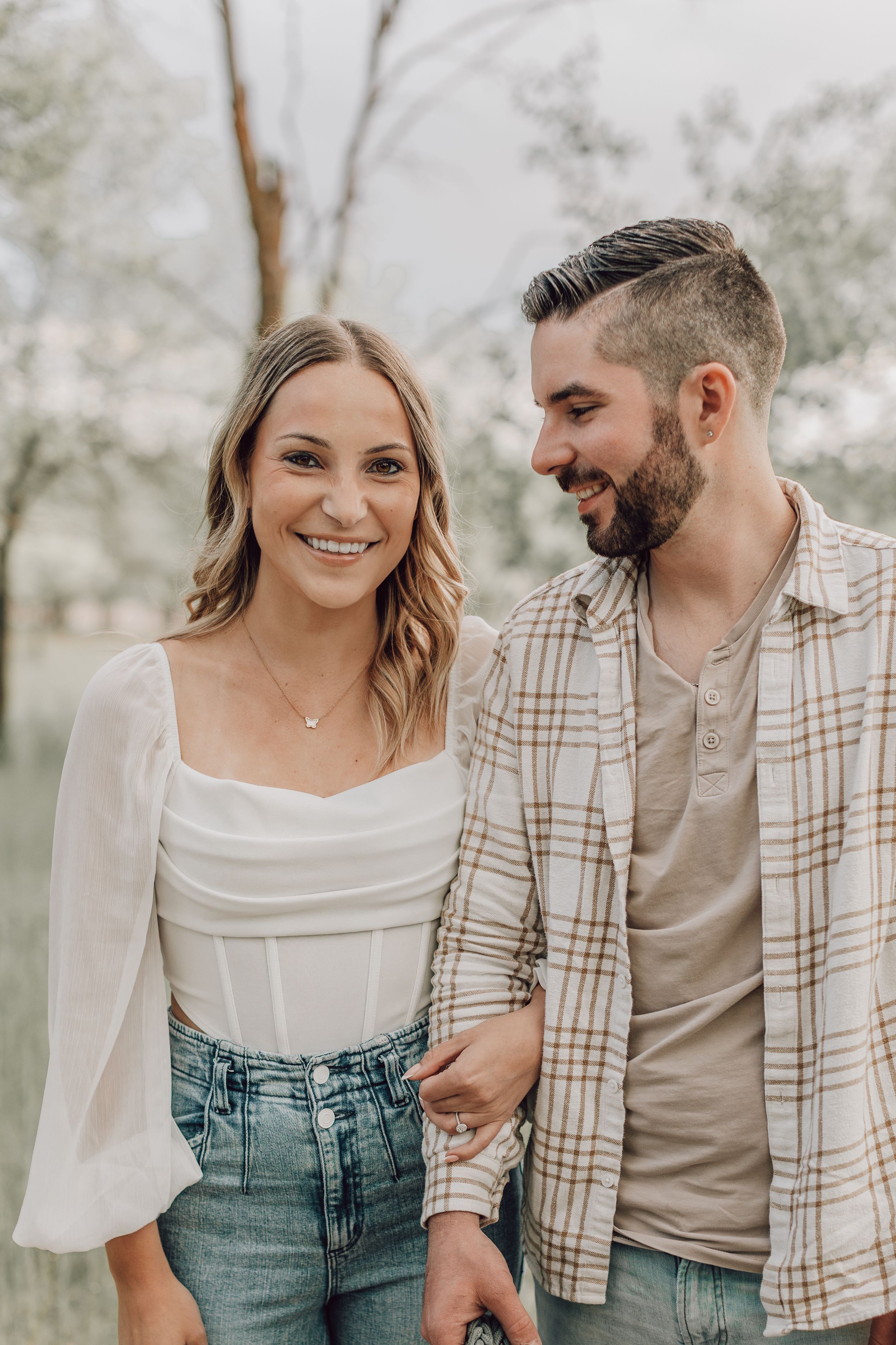 A smiling couple walks arm in arm outdoors, wearing casual outfits in a park setting.