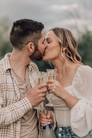 An engaged couple kisses while clinking champagne glasses to celebrate their engagement.