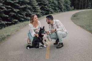 A happy couple crouches on a road with their black and white dog in a forest setting.