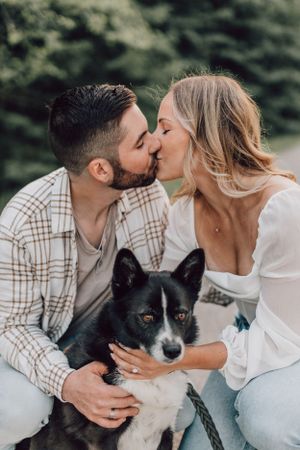 An engaged couple kisses while holding their black and white dog during an outdoor photoshoot.