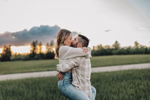A man lifts and kisses a woman in a green field at sunset during an engagement session.