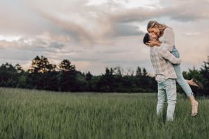 A couple shares a romantic lift and kiss in a lush green field under a dramatic cloudy sky.