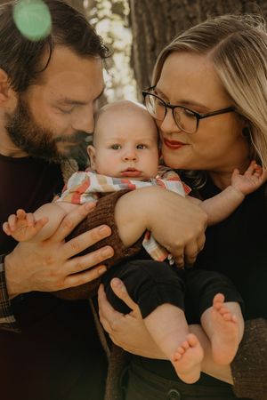 Loving parents tenderly holding and kissing their infant baby boy outdoors near a tree.