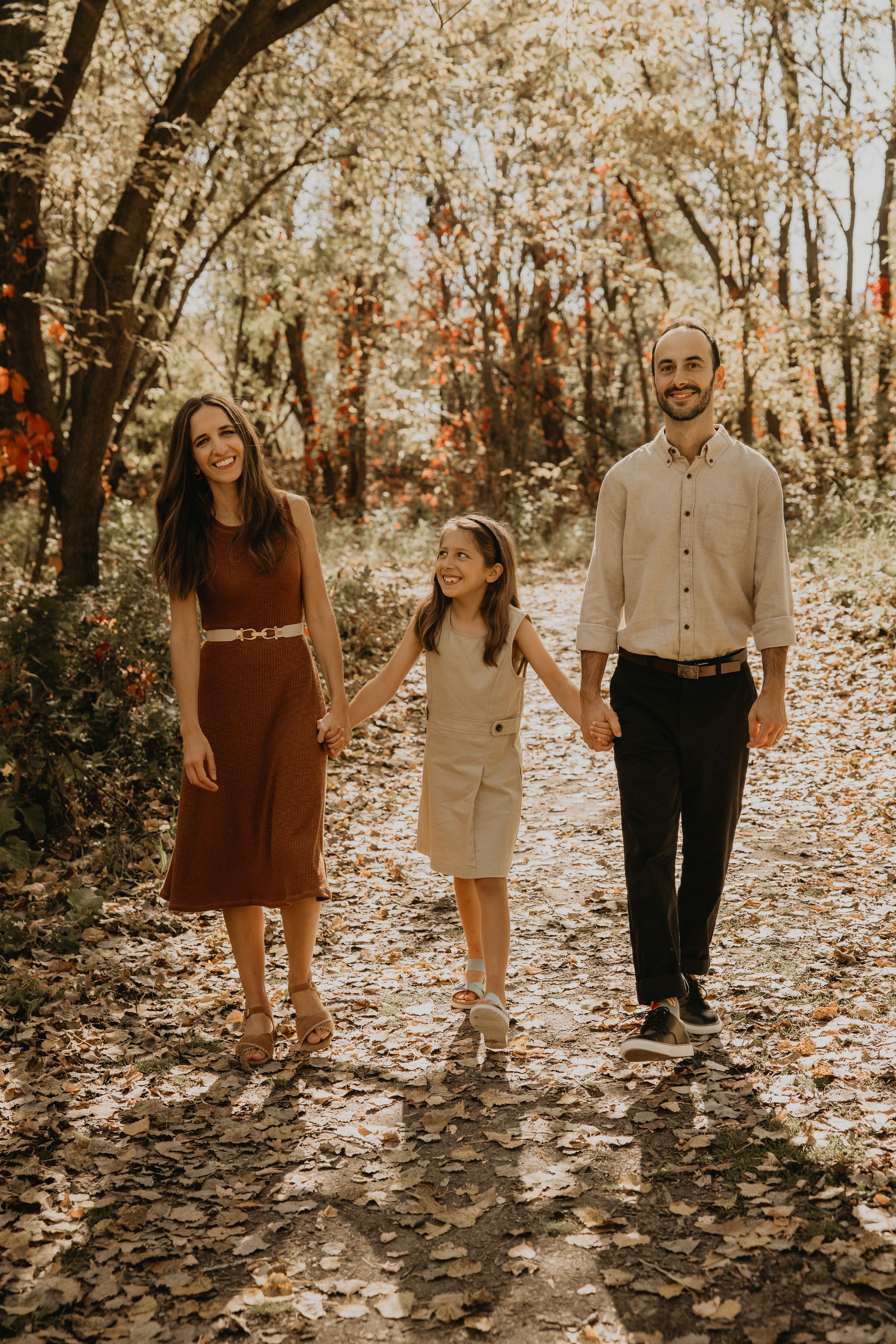Happy family of three walking hand-in-hand along a leaf-covered path in an autumn forest.