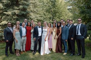 Large wedding group photo of guests and couple in formal attire posing outdoors under green trees.