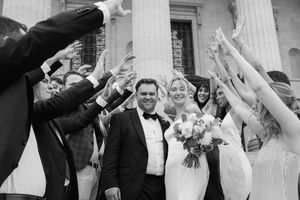 Black and white photo of a bride and groom celebrating with guests raising their hands outside a grand building.