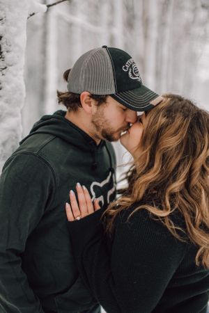 Couple sharing a kiss in snowy winter forest during engagement photo session.