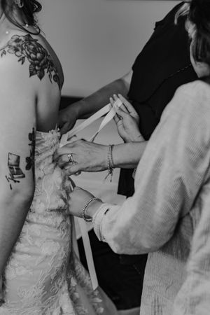 Black and white photo of helpers lacing up tattooed bride's wedding dress corset back.