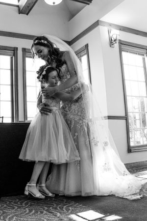 Black and white photo of bride in lace gown hugging young flower girl indoors.