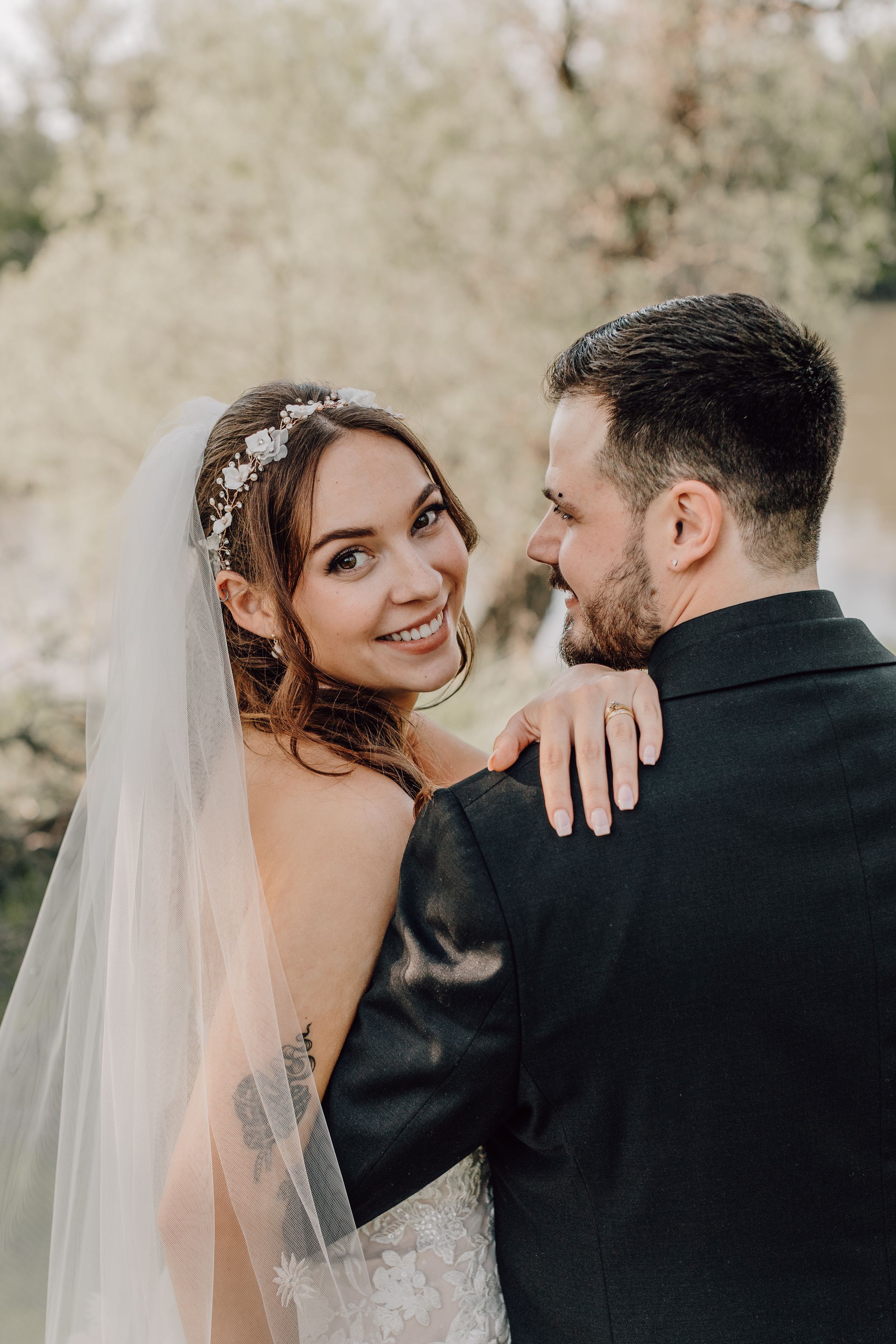 Smiling bride in lace gown and veil embracing groom outdoors during wedding portraits.