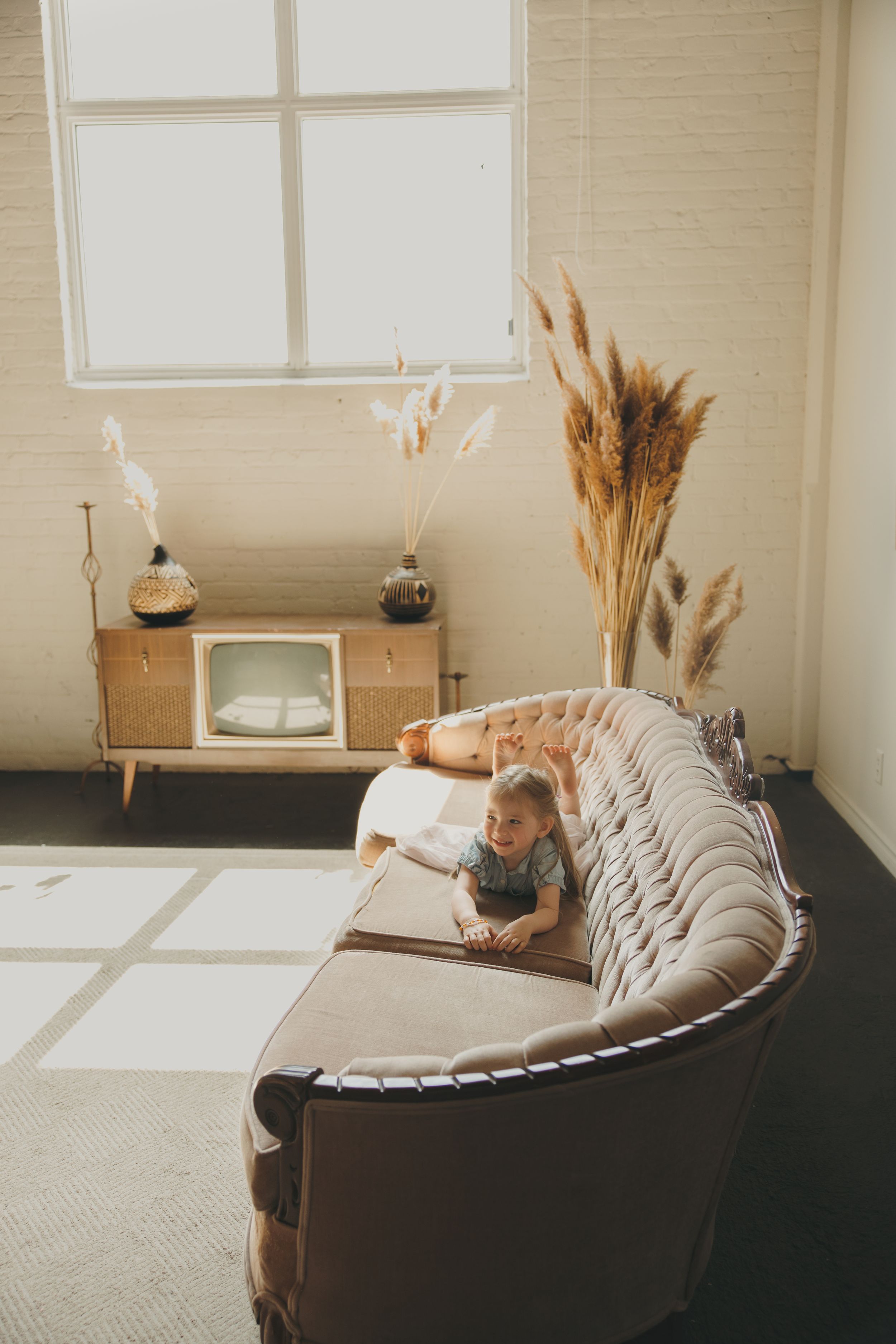 A baby crawling on a tufted beige sofa in a bright, modern living room with pampas grass decor.