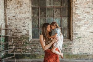 A mother tenderly kisses her young daughter in front of a rustic brick wall indoors.
