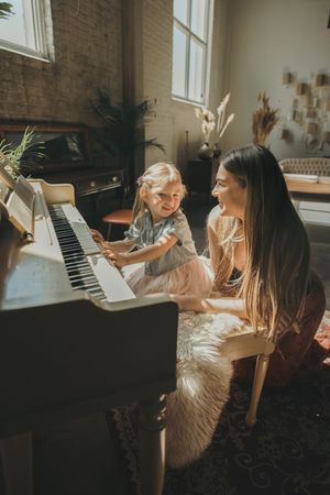 A mother and young daughter share a joyful moment playing piano together in a sunlit room.