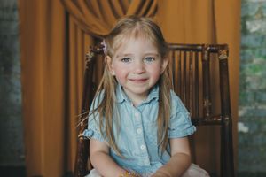 Adorable blonde girl with pigtails smiling while sitting in a wooden chair indoors.