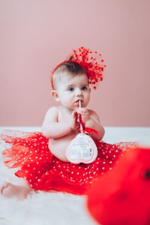 Adorable baby girl in red tutu skirt and bow headband sitting on white fur rug with Valentine gift.