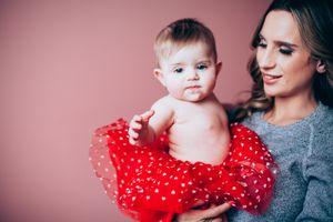 Young mother in gray sweater lovingly holds baby girl dressed in red polka dot tutu against pink background.