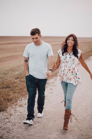 Couple holding hands and walking on a rural dirt road through open fields.