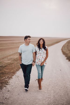 Smiling couple walking hand in hand along a long straight dirt road in open farmland.