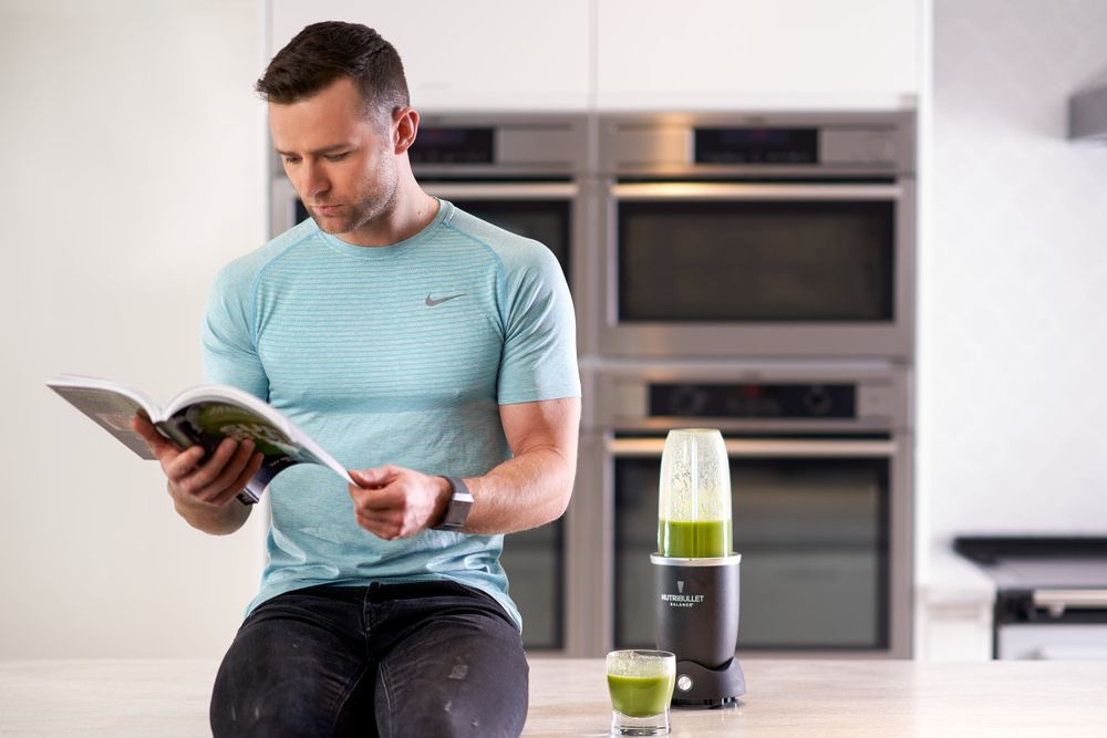 Harry Judd sitting on a kitchen counter, reading a book with a blender next to him