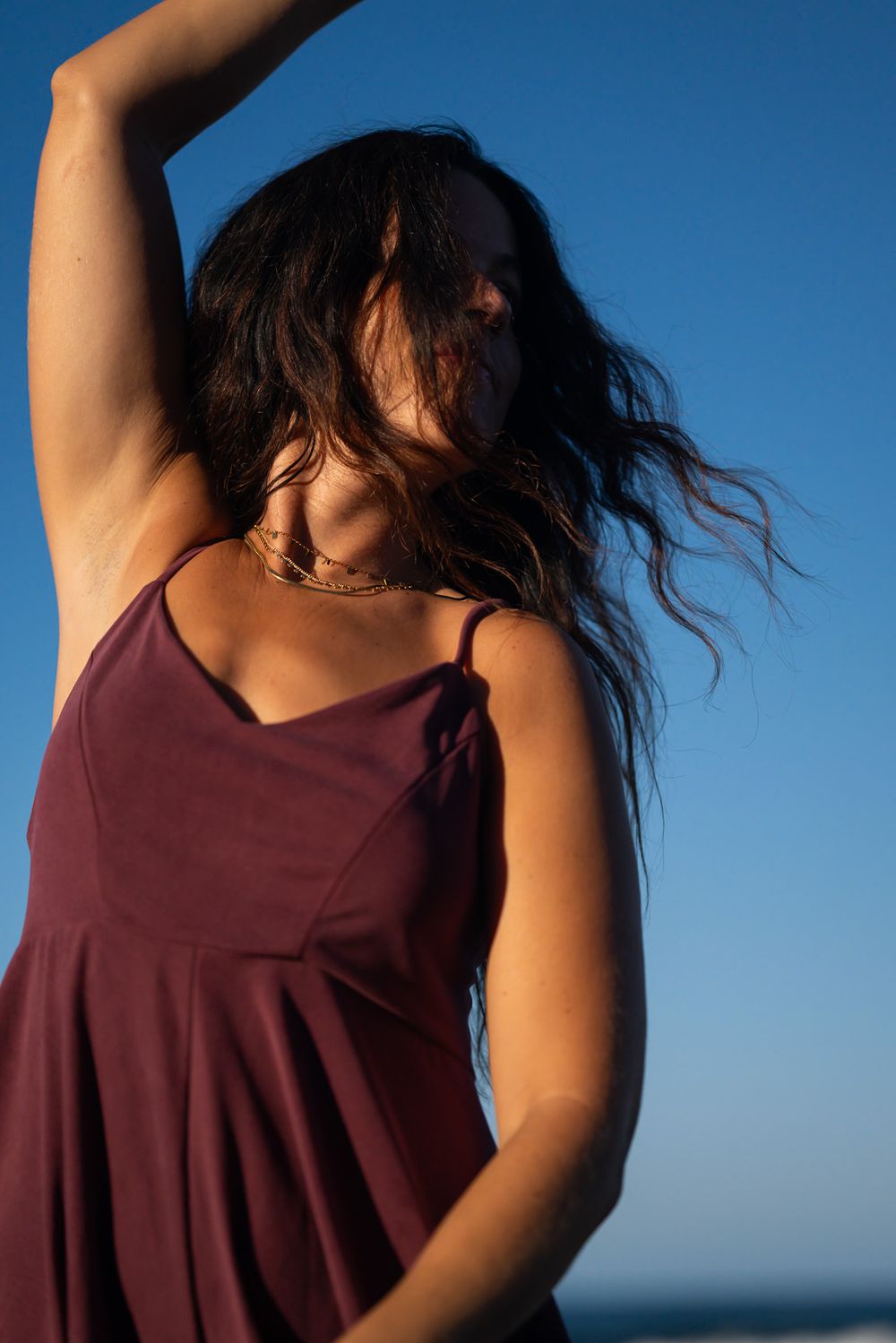 Woman in burgundy dress dancing with flowing hair against bright blue sky at beach.
