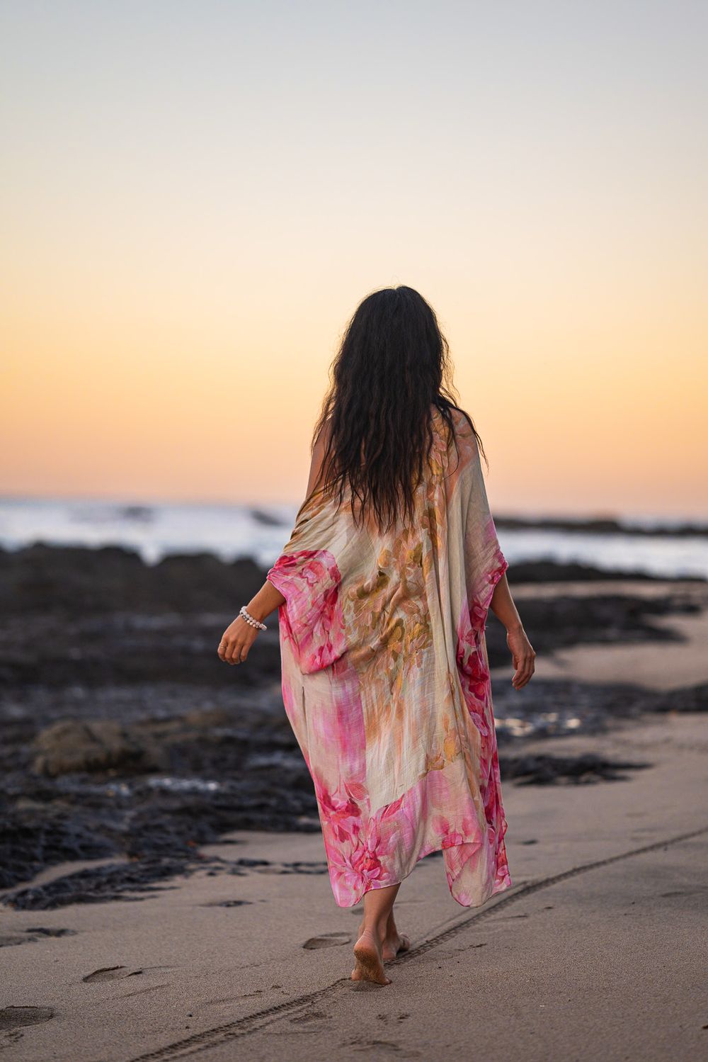 Person walking on beach at sunset wearing flowing pink floral kimono.