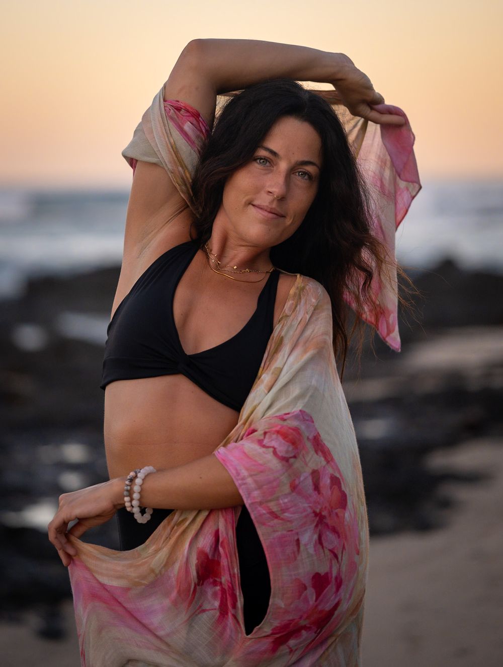 Beach portrait with flowing pink floral wrap against rocky coastal sunset.