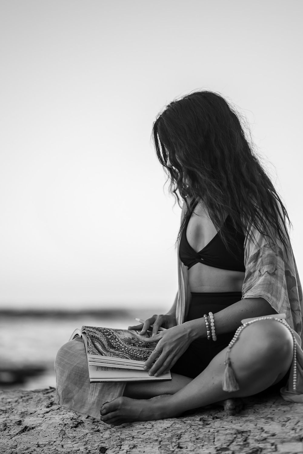Black and white photo of person reading book on beach wearing black swimwear.
