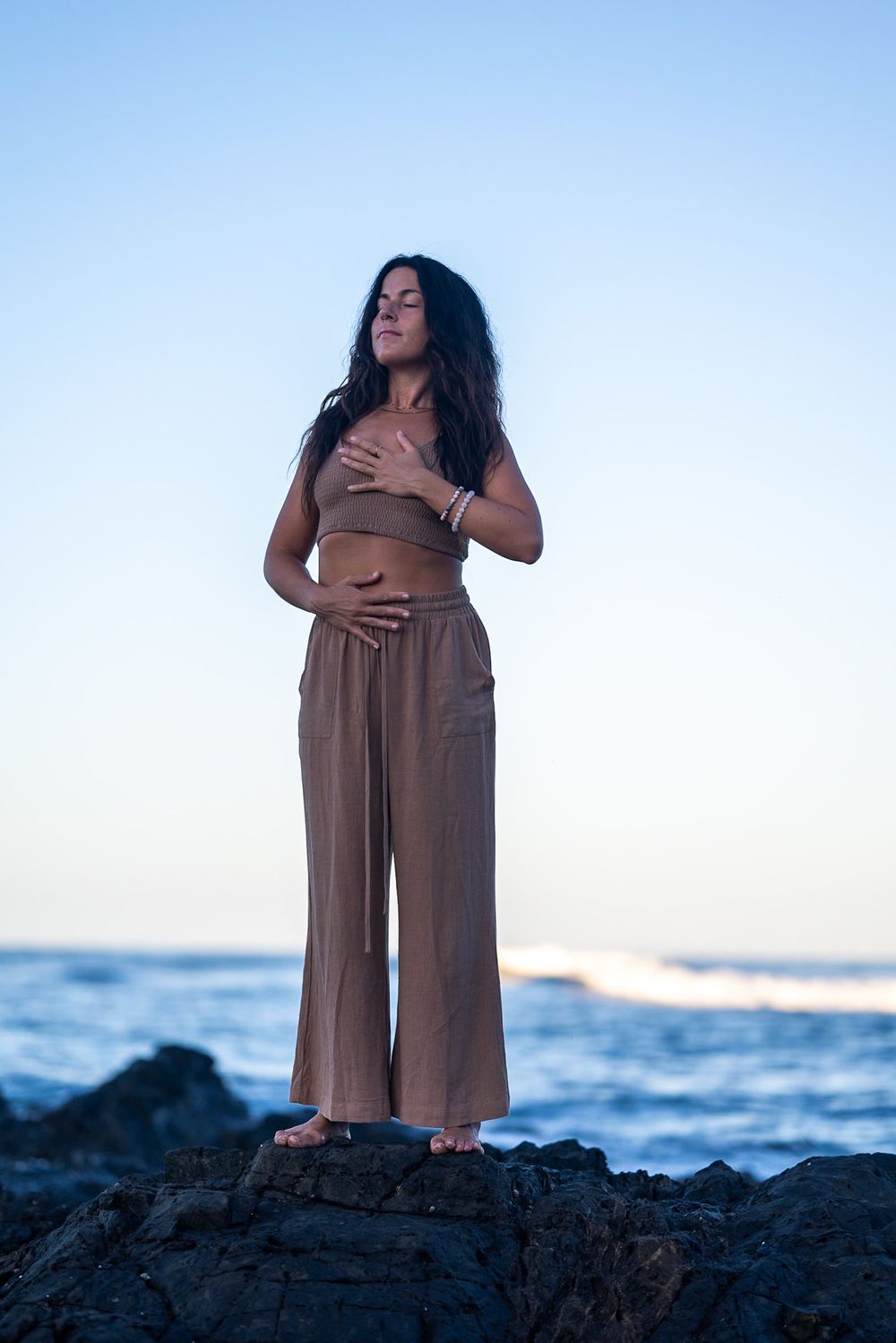 Silhouette standing on coastal rocks at sunset in flowing brown outfit.