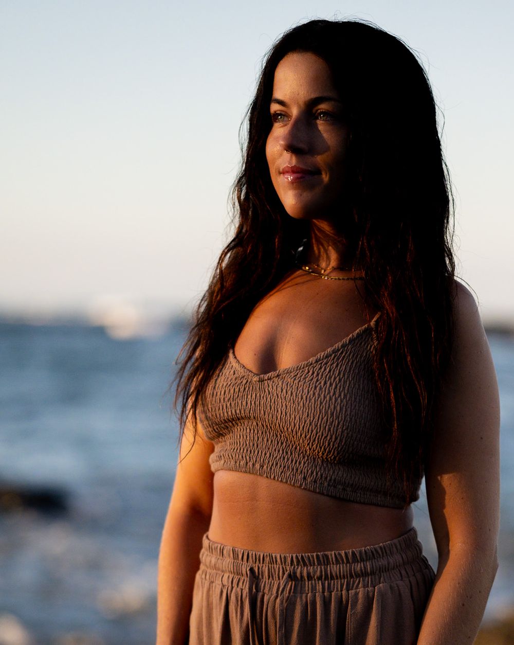 Woman in knit crop top set poses against ocean backdrop during golden hour.