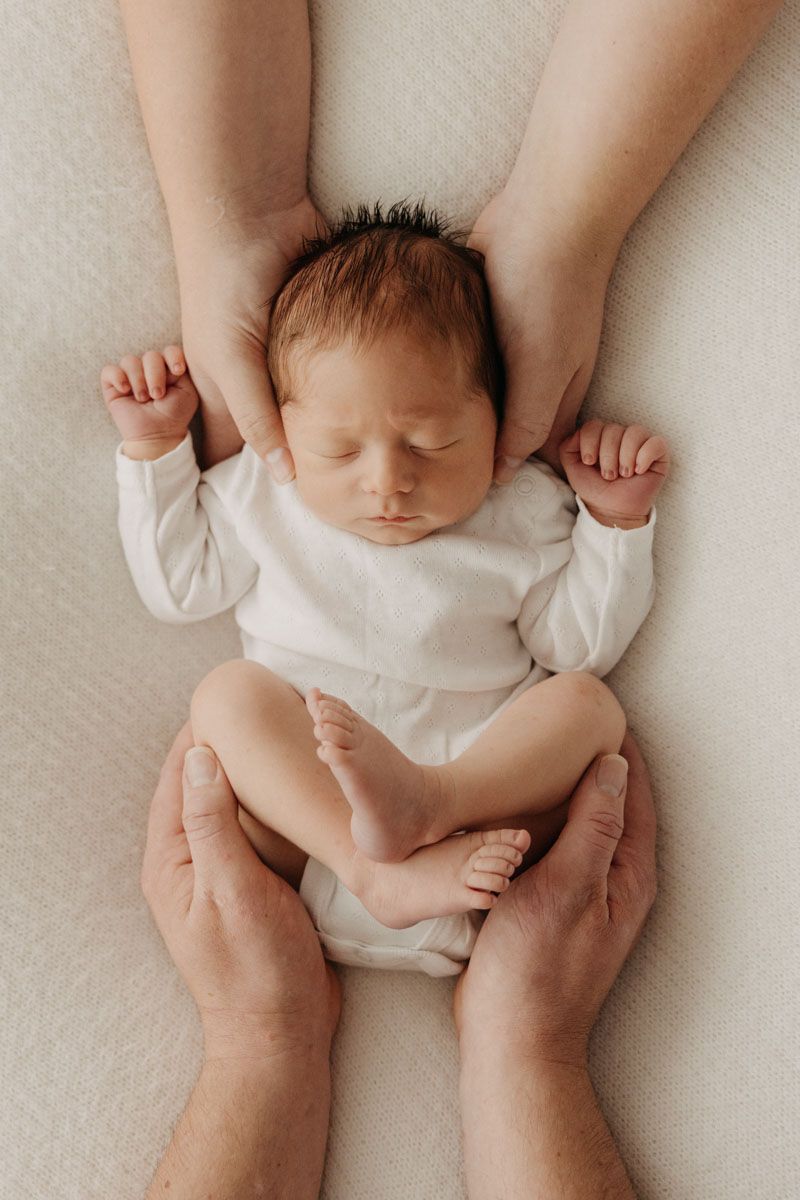 Gentle hands cradle sleeping newborn baby wearing white onesie on soft white fabric.