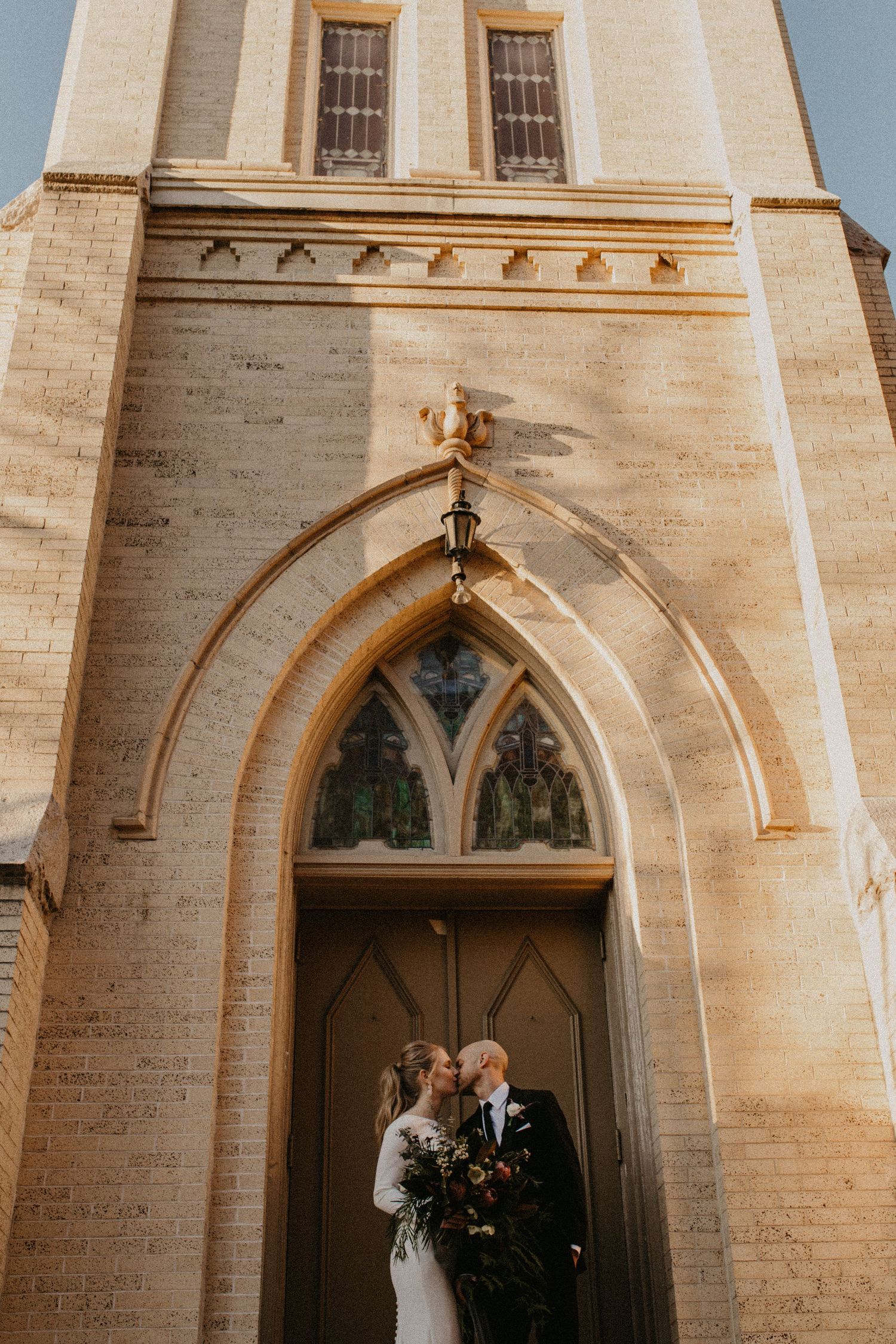 A romantic wedding moment captured in front of a gothic church doorway with arched architecture and warm beige stonework.