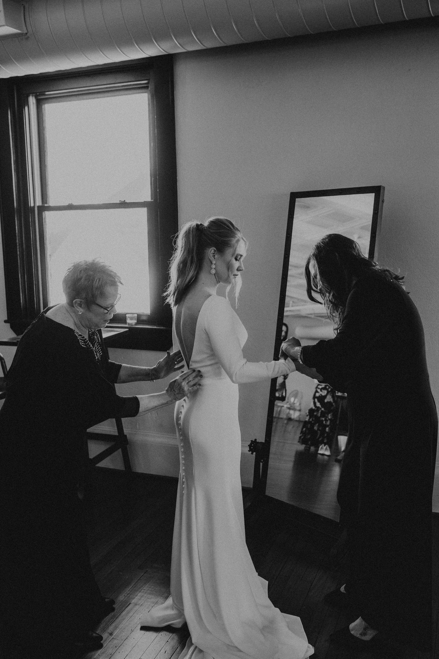 Black and white wedding photo showing final dress adjustments being made to a bride wearing an elegant fitted gown.