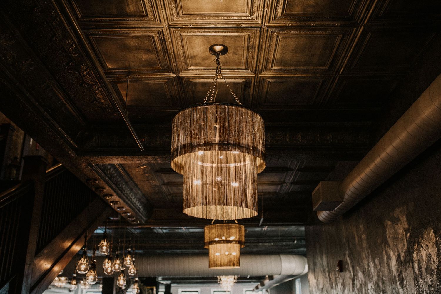 Crystal chandeliers with tiered designs hang from an ornate tin ceiling in a dimly lit industrial-style space.