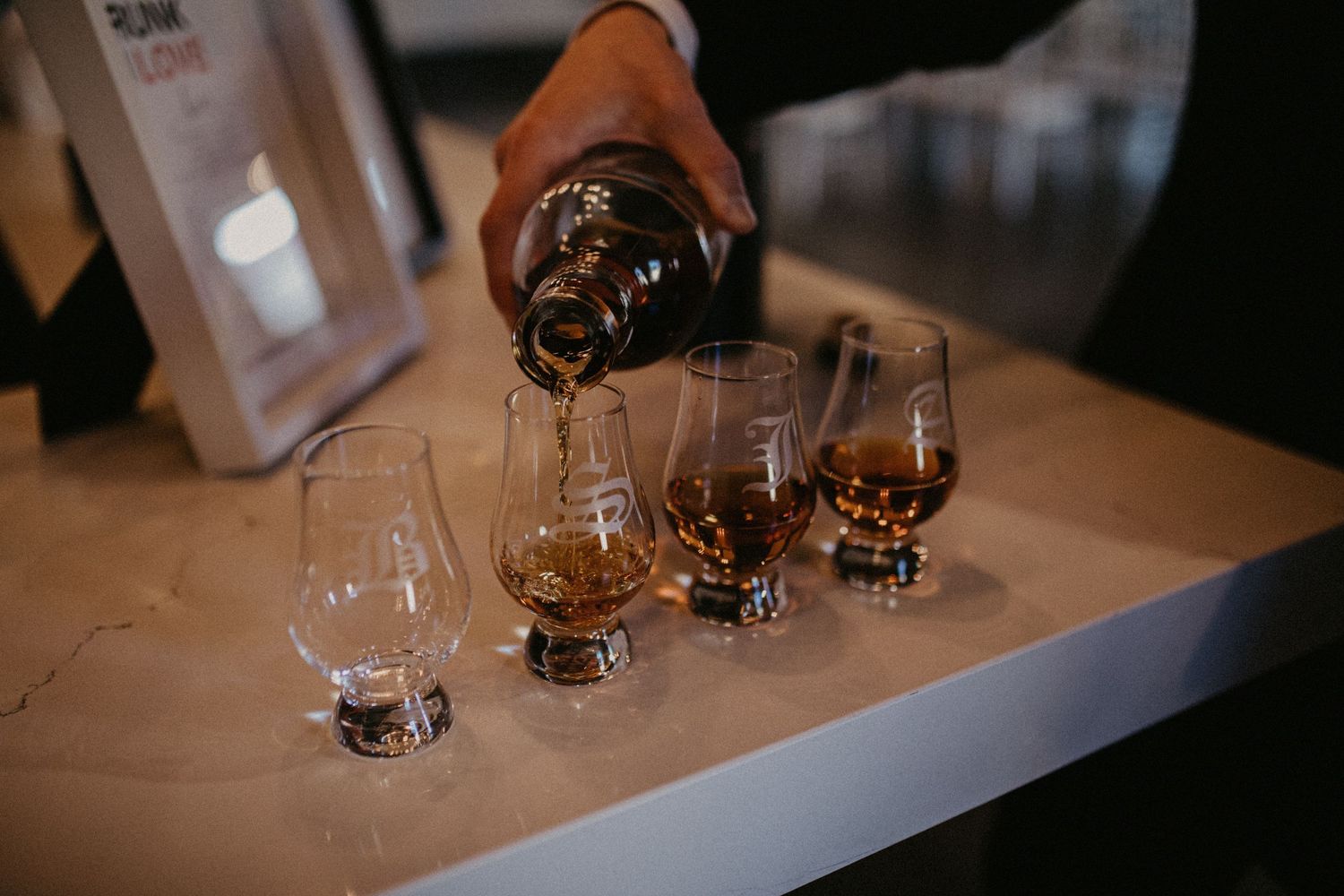 A bottle of dark liquor being poured into three whiskey tasting glasses on a wooden table in moody lighting.